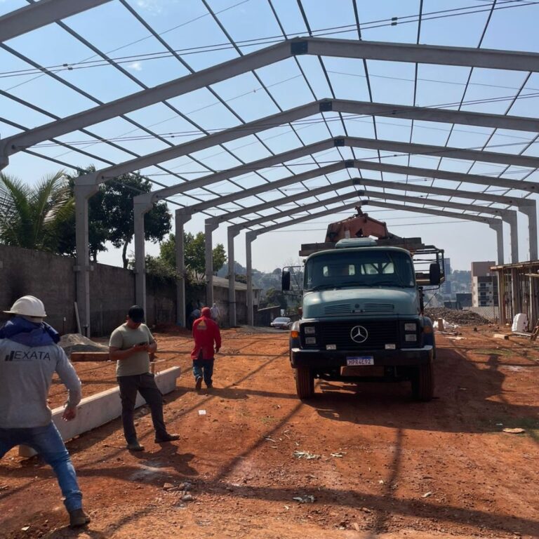 Pilares e vigas de concreto pré-moldado formando a estrutura de um galpão industrial em fase inicial de construção, com fundações e área de obra ao fundo.